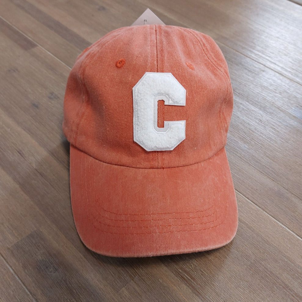 Casquette Ponteau est en tissus uni délavé de forme base Ball coloris corail, réglable par l'arrière elle a un C majuscule en moumoute blanche sur le devant