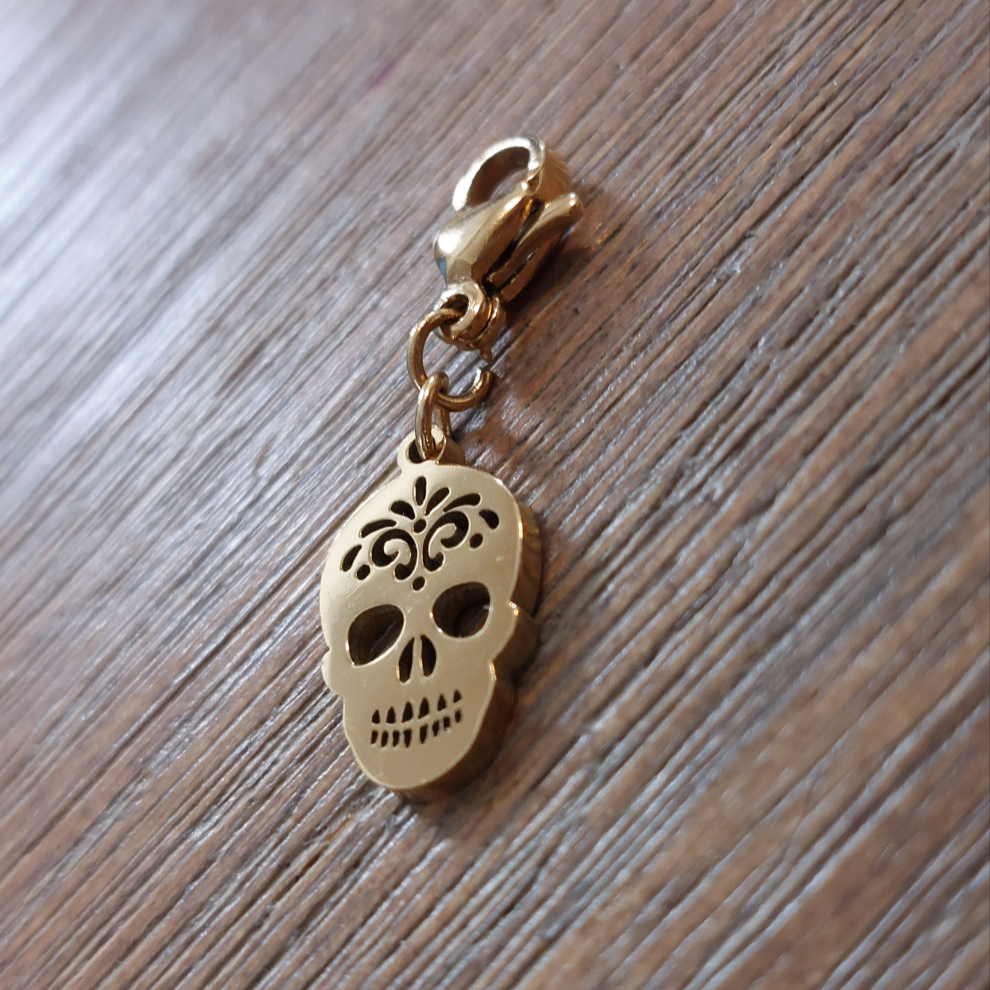 charm ou breloque avec un petit fermoir pour ajouter à un collier où à un bracelet en accumulation personnalisé. il represente une tête de mort doré et ajoureé en acier inoxydable sur carry le rouet