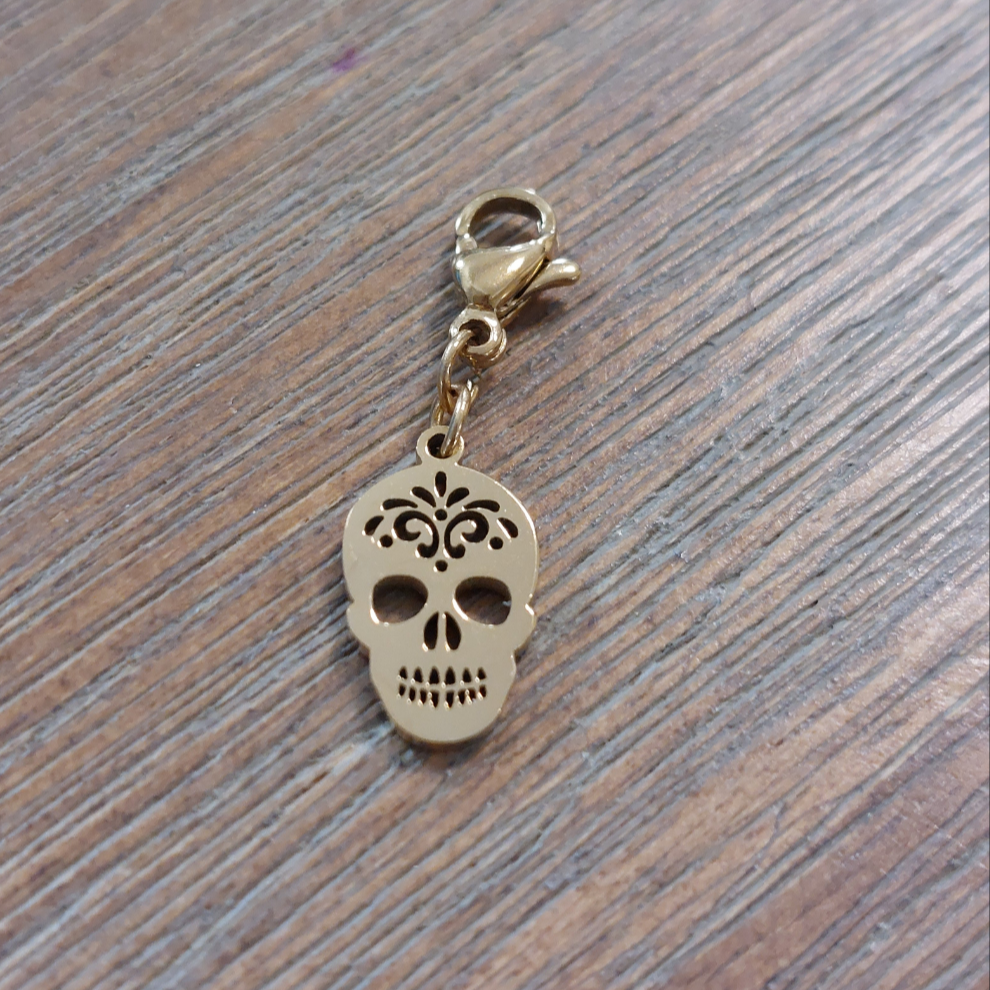 charm ou breloque avec un petit fermoir pour ajouter à un collier où à un bracelet en accumulation personnalisé. il represente une tête de mort doré et ajoureé en acier inoxydable sur aix en provence