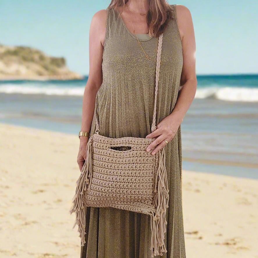 sac en maccramé porté en bandoulière. Il possède des franges sur toute la longueur de chaque cotés, une poignée rectangle decoupée dans la matère sur le haut du cas et il est en coloris camel sur mar gnane