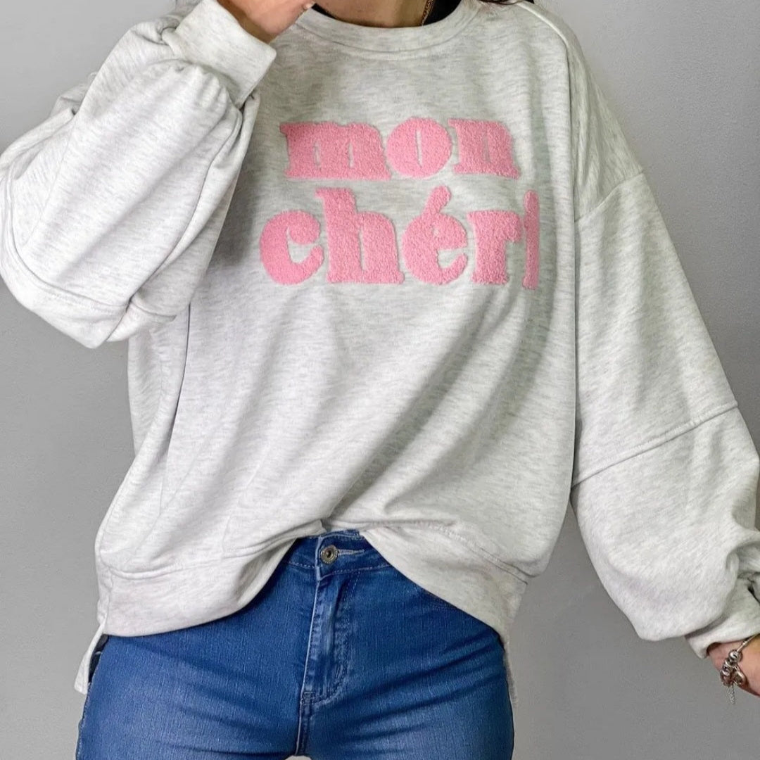 Sweat manches longues finis par un poigné a bord cotelé et porté par un mannequin sur un jean. col rond et l'on voit devant une grosse incription MON CHERI en bouclette rose sur carry le rouet