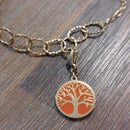 charm ou breloque avec un petit fermoir pour ajouter à un collier où à un bracelet en accumulation personnalisé. il represente une pastille ronde motif arbre de vie sur fond corail  doré en acier inoxydable monté sur un bracelet anneaux martelés sur aix en provence