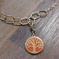 charm ou breloque avec un petit fermoir pour ajouter à un collier où à un bracelet en accumulation personnalisé. il represente une pastille ronde motif arbre de vie sur fond corail  doré en acier inoxydable monté sur un bracelet anneaux martelés sur aix en provence