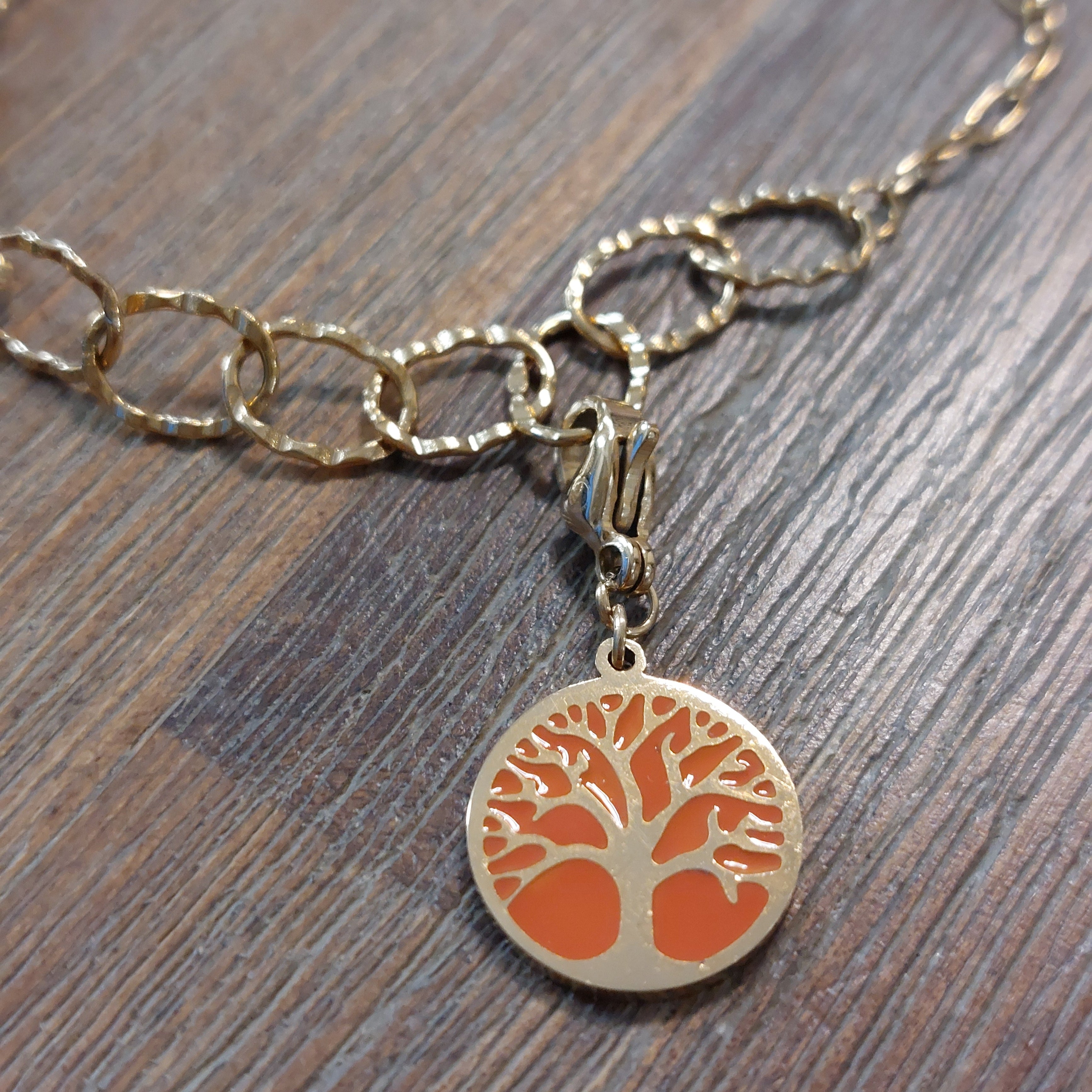 charm ou breloque avec un petit fermoir pour ajouter à un collier où à un bracelet en accumulation personnalisé. il represente une pastille ronde motif arbre de vie sur fond corail  doré en acier inoxydable monté sur un bracelet anneaux martelés sur aix en provence