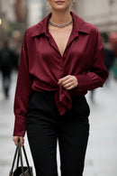 Chemise satinée Gloria bordeaux portée nouée face – col chemise ouvert, manches longues manchettes boutonnées, pantalon noir taille haute, collier chaîne argent, sac tote noir, style urbain chic
