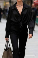 Chemise satinée Gloria noir portée nouée face – col chemise ouvert, manches longues manchettes boutonnées, pantalon noir taille haute, total look noir collier chaîne argent, sac tote kaki

