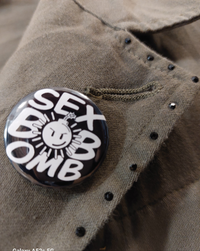 Gros plan sur le pin's rond vintage inscription blanc sur noir en lettre majuscule "SEX BOB OMB" le o de bob remplacé par une bomb smiley pas content