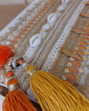Gros plan des ornements de la Pochette Nandi – tresses dorées, coquillages, sequins orange et glands bicolores orange et moutarde
