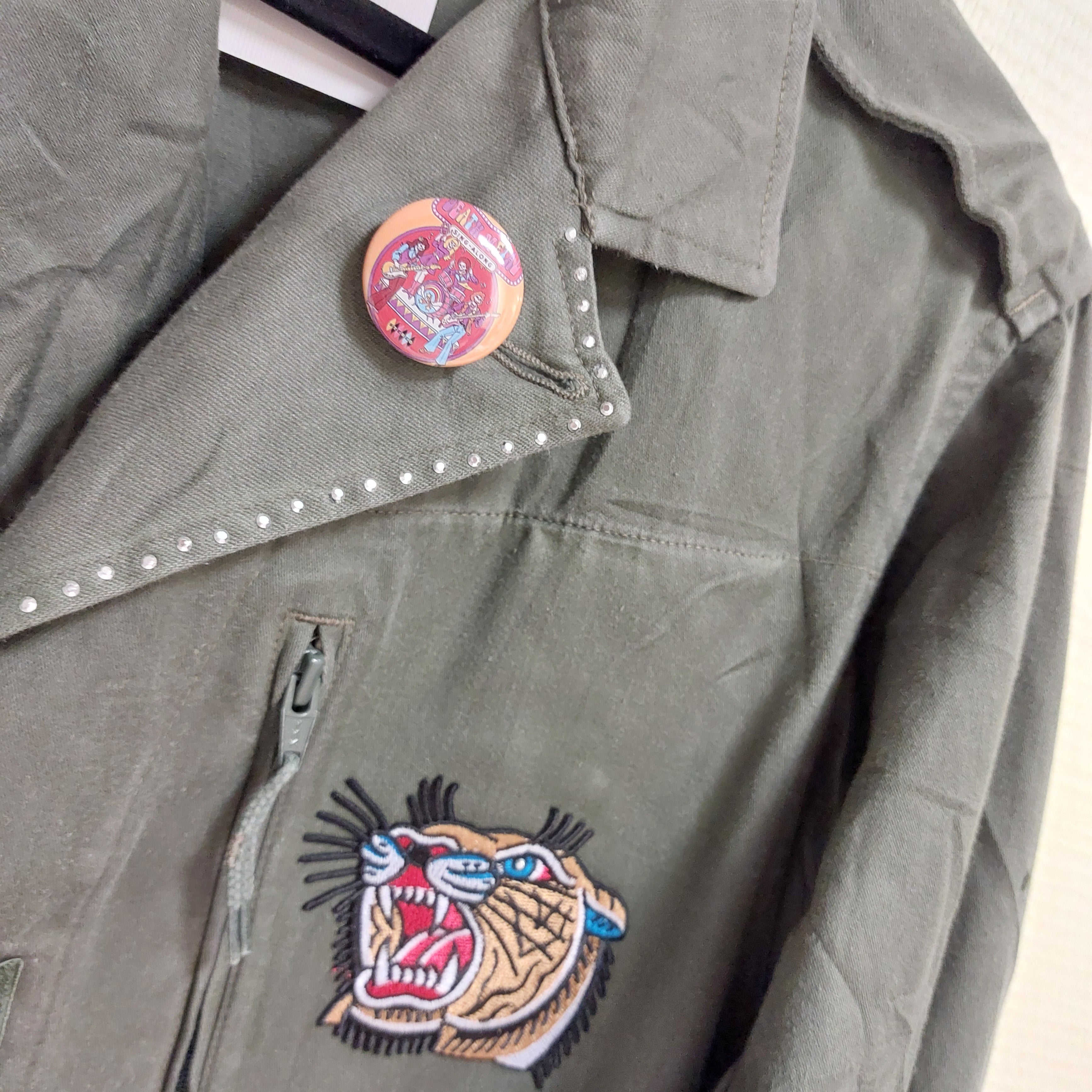 veritable veste mlitaire unie kaki. elle est customizée avec un patch tete de tigre en couleur sur poitrine gauche, elle a un badge rond sur le revers gauche du col representant un groupe de metal    sur carry le rouet