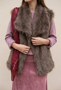 Gilet sans manches Loren choco en fausse fourrure à poils longs, porté sur un pull rose et sa jupe en satin assortie.