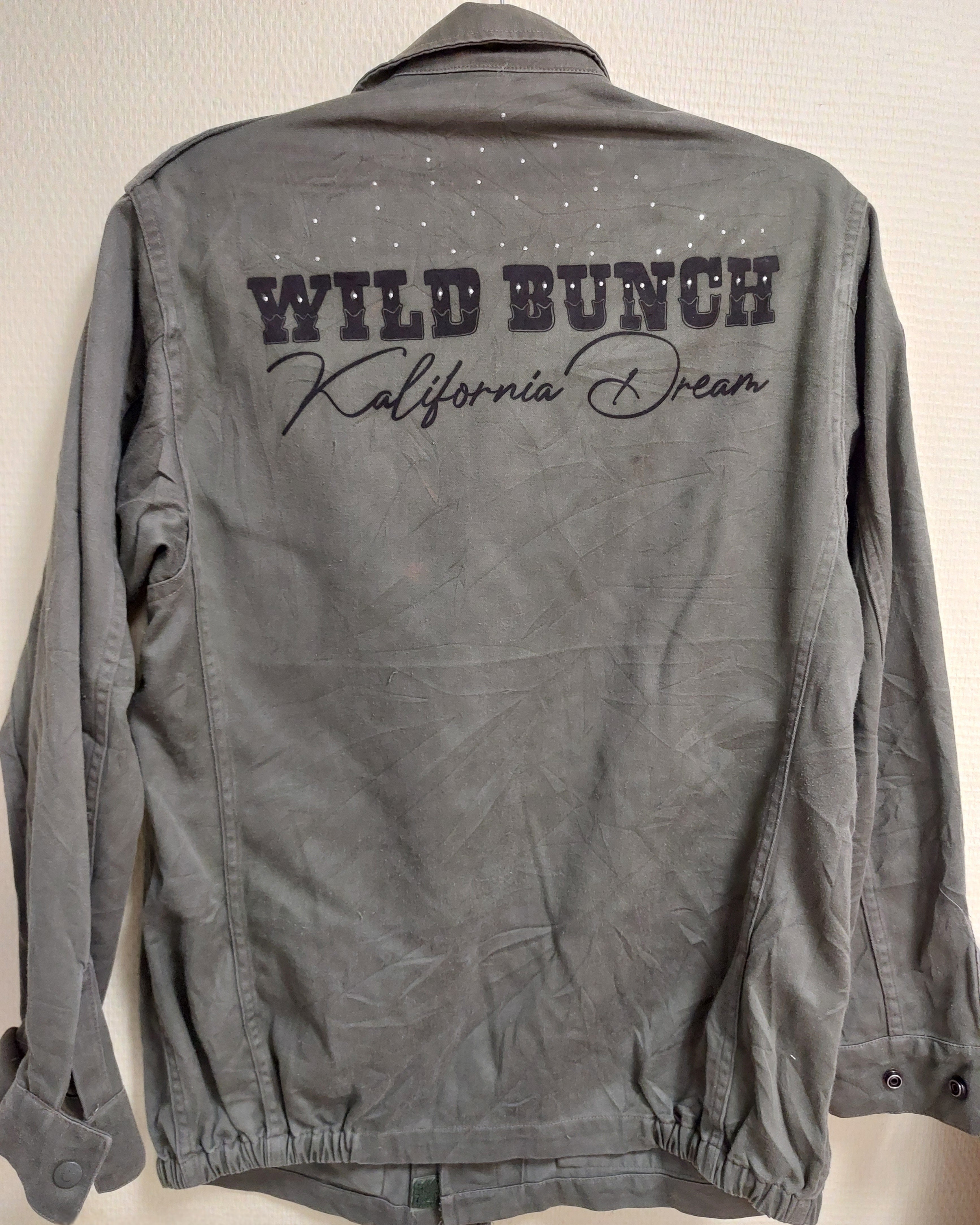 veritable veste mlitaire unie kaki longueur sous fesses vue de dos. elle est customizée avec une inscription sur tout le haut en majuscule dans un style western "WILD BUNCH Kalionia Dream" et une série de petit clou acier par çi par là . sur aix en provence