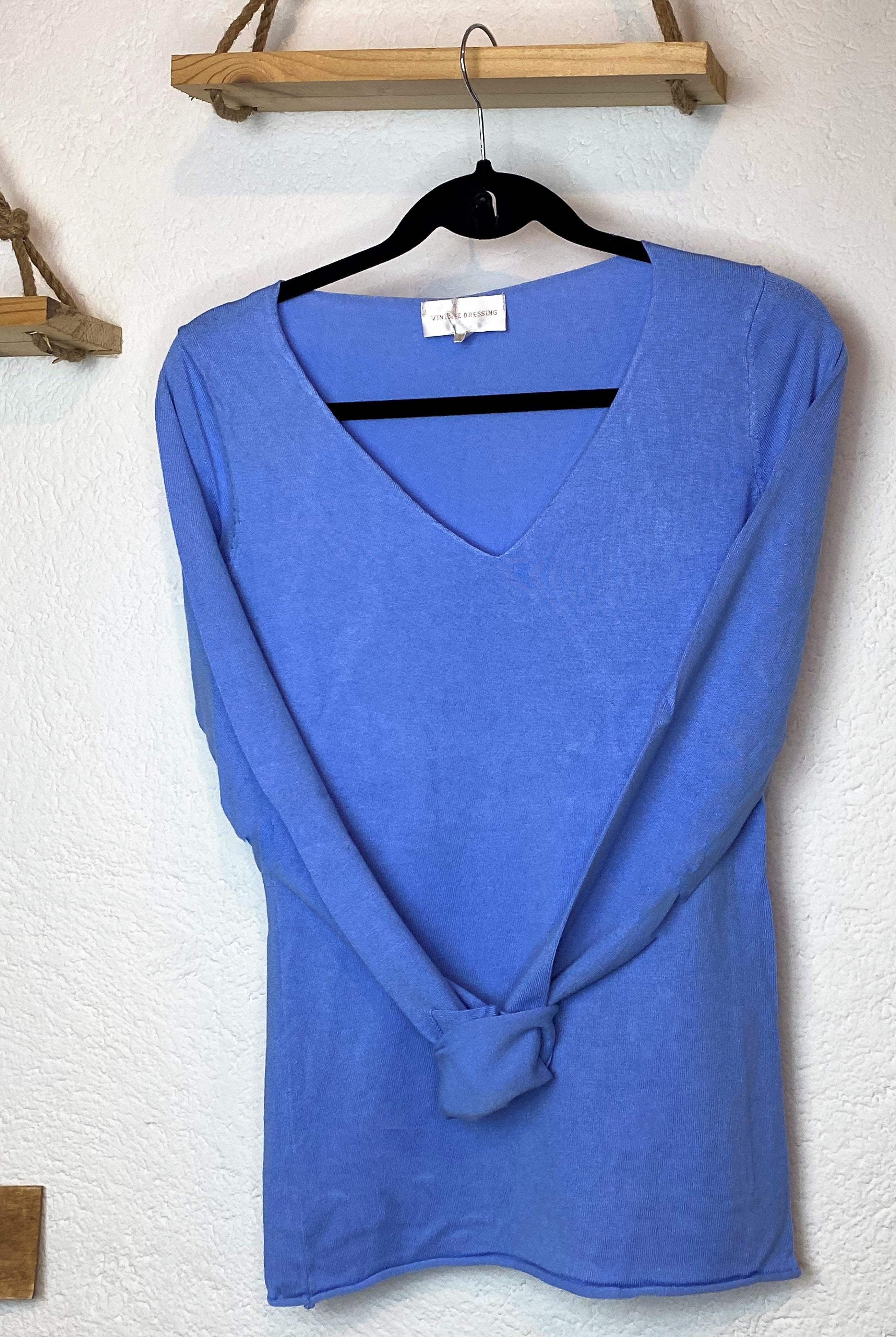 Un Pull Marlène bleu d'Ineva sur cintre noir est accroché contre un mur texturé blanc. La chemise semble être soigneusement exposée en mode, et il y a deux étagères en bois avec des supports en corde visibles en arrière-plan.