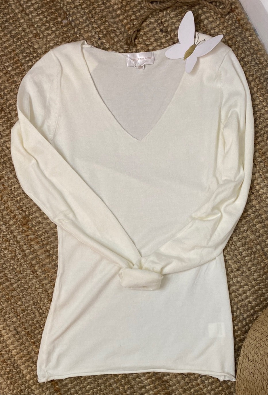 Une chemise blanche à manches longues et col en V est soigneusement disposée sur une surface texturée beige. Un papillon en papier blanc est attaché au col, apportant une touche délicate et fantaisiste à la présentation, parfait pour mettre en valeur des accessoires féminins élégants. Le Pull Marlène d'Ineva ajoute une élégance sophistiquée à la scène.